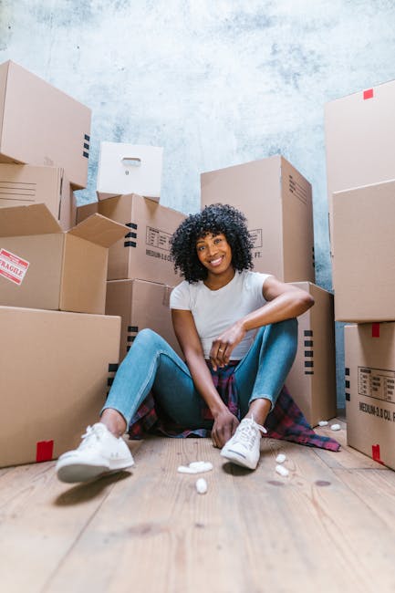 A young woman with curly black hair and a bright smile is sitting on a wooden floor amidst numerous packed cardboard boxes of various sizes, some with black and red markings, all sealed with packing t