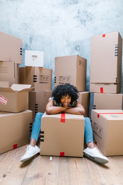 A young woman with curly black hair and a bright smile is sitting on a wooden floor amidst numerous packed cardboard boxes of various sizes, some with black and red markings, all sealed with packing t